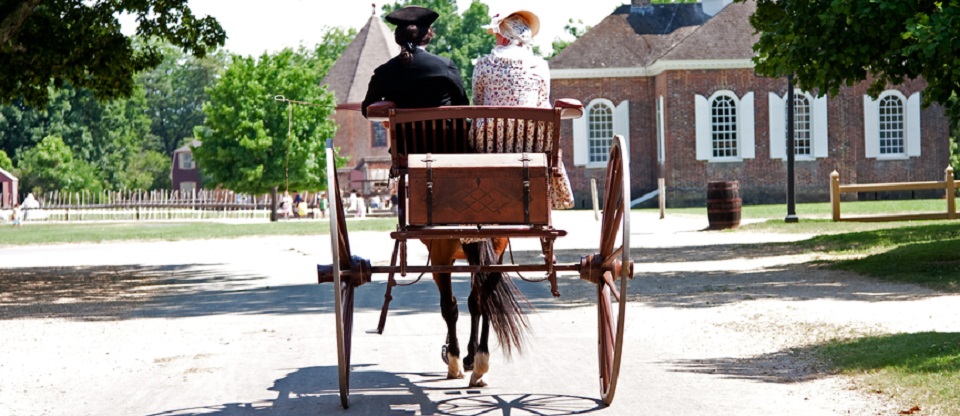 Rear view of a historical couple of a man and a woman dressed in Colonial American style on a brown carriage with treasure chest pulled by a horse in Colonial Williamsburg, Virginia